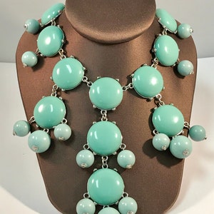 May include: A turquoise statement necklace with large round beads and smaller round beads hanging from the larger beads. The necklace is made of metal and has a silver finish.