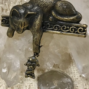 May include: A gold-toned metal brooch of a cat hanging from a ledge, with a small mouse dangling from its paw.