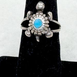 May include: A silver ring featuring a turtle-shaped centerpiece. The turtle has a textured shell design with a small, round, bright blue stone set in the center. The ring band is thin and splits to connect to the sides of the turtle figure.