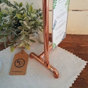 May include: Copper pipe stand with a sign holder for a wedding menu. The sign reads "D & C Wedding Menu". The stand is on a white lace tablecloth with a brown tag that says "Rainey Lane Design".