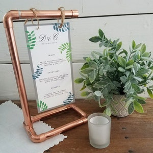 Puede incluir: Un soporte de tubería de cobre con un letrero que dice "D&C Wedding Menu". El letrero tiene un diseño de hojas y enumera los elementos del menú de la boda. Una pequeña planta en maceta y una vela están sobre la mesa.