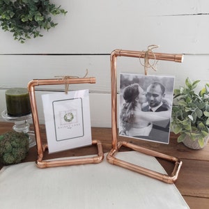 Może przedstawiać: Dwa ramki na zdjęcia z miedzianych rur z rustykalnym sznurkowym wieszakiem. Ramka po lewej stronie zawiera wizytówkę z tekstem "Rainey Lane Designs". Ramka po prawej stronie zawiera czarno-białe zdjęcie pary.