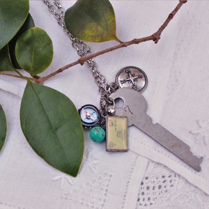 Puede incluir: Un collar plateado con un colgante de llave grande, un dije de brújula y un pequeño dije rectangular. El collar también presenta una cuenta turquesa y un dije circular con un diseño de rosa de los vientos. Hojas verdes y una rama en el fondo.