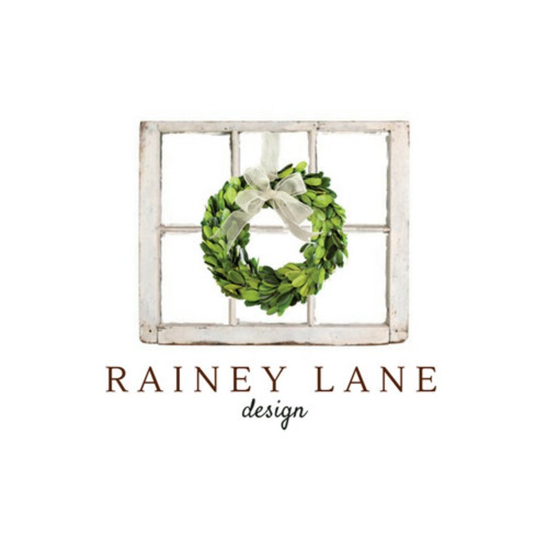 May include: A white window frame with a green boxwood wreath hanging from a white ribbon. The text "Rainey Lane design" is below the window frame.