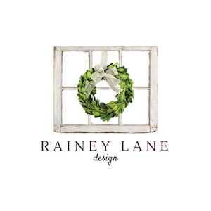 Op de afbeelding: Een wit raamkozijn met een groene buxuskrans die aan een wit lint hangt. De tekst "Rainey Lane design" staat onder het raamkozijn.