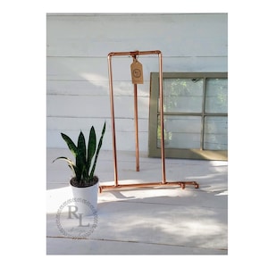 May include: A copper pipe plant stand with a small potted plant. The stand is made of copper pipes and has a simple design. The stand is on a white wooden surface.