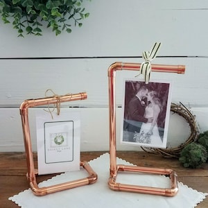 Op de afbeelding: Twee fotohouders van koperen pijp met een witte achtergrond. De houder rechts heeft een zwart-witfoto van een stel. De houder links heeft een kaart met de tekst "Rainey Lane Designs".