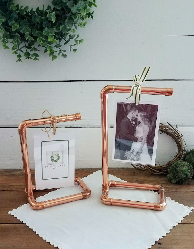 Op de afbeelding: Twee fotohouders van koperen pijp met een witte achtergrond. De houder rechts heeft een zwart-witfoto van een stel. De houder links heeft een kaart met de tekst "Rainey Lane Designs".