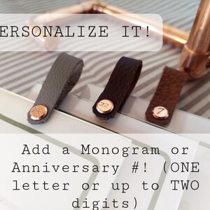 Op de afbeelding: Drie leren riemen met metalen accenten. De riemen zijn grijs, bruin en zwart. Elke riem heeft een ander nummer of een andere letter erop. De tekst "PERSONALIZE IT!" staat bovenaan de afbeelding. De tekst "Add a Monogram or Anniversary #! (ONE letter or up to TWO digits)" staat onderaan de afbeelding.