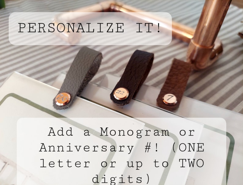 Op de afbeelding: Drie leren riemen met metalen accenten. De riemen zijn grijs, bruin en zwart. Elke riem heeft een ander nummer of een andere letter erop. De tekst "PERSONALIZE IT!" staat bovenaan de afbeelding. De tekst "Add a Monogram or Anniversary #! (ONE letter or up to TWO digits)" staat onderaan de afbeelding.