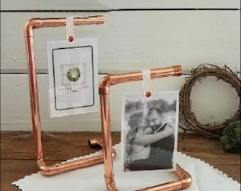 May include: Two copper pipe picture frames with twine and a small wooden bead hanging from the top. The frames are holding photos. The frame on the left has a photo with the text "Raines Lane Design". The frame on the right has a photo of a couple embracing.