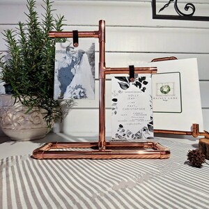 May include: Copper pipe picture stand with three photo slots. The stand is holding a wedding invitation with a floral design and two photos. The invitation reads "Holly Jean and Paul Christopher".