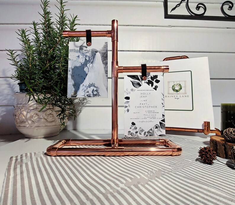 May include: Copper pipe picture stand with three photo slots. The stand is holding a wedding invitation with a floral design and two photos. The invitation reads "Holly Jean and Paul Christopher".