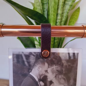Op de afbeelding: Een fotohouder van koperen pijp met een bruine leren band en een metalen klinknagel. Een zwart-witfoto van een stel wordt in de houder weergegeven.