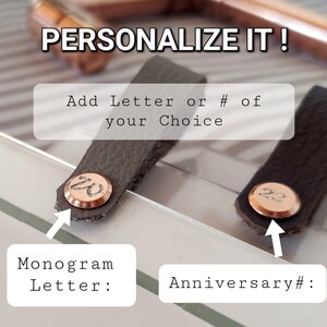 May include: Two gray leather tabs with a copper colored metal disc. The left tab has the letter "W" engraved on the disc. The right tab has the number "22" engraved on the disc. The text "PERSONALIZE IT!" is above the tabs. The text "Add Letter or # of your Choice" is below the tabs. The text "Monogram Letter:" is below the left tab. The text "Anniversary #:" is below the right tab.