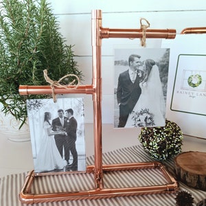 May include: Copper pipe photo display stand with two black and white wedding photos and a card with the text "Rainey Lane Design".