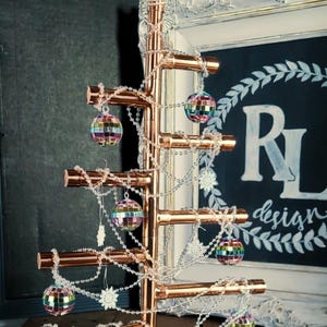 Op de afbeelding: Een kerstboom van koperen pijpen, versierd met regenboogkleurige discoballen en een witte parelketting. De boom is gekroond met een witte ster en staat op een houten oppervlak.