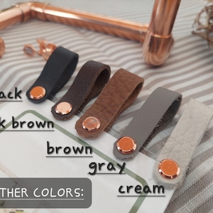 May include: Leather straps in black, dark brown, brown, gray, and cream colors. The straps have a copper rivet on each end.  LEATHER COLORS: is written on the surface below the straps.