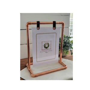 May include: Copper pipe picture frame with black leather straps holding a clear acrylic sheet. The frame holds a card with a wreath design and the text "RAINEY LANE design". The frame is on a white surface.