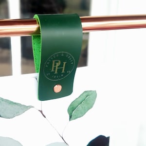 May include: A dark green leather hanger with a copper rivet and a logo. The hanger is attached to a copper rod. The interior of the hanger is a lighter green. The logo reads "Payver & Holly".