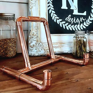 May include: A copper pipe book stand with a rustic design. The stand is made of copper pipes and fittings and has a simple, industrial look. It is perfect for displaying books, magazines, or other items.