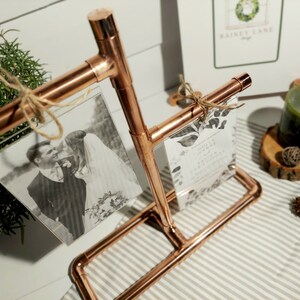 May include: Copper pipe picture frame with two photos and a wedding invitation. The frame is on a striped surface with a candle and other decorations.