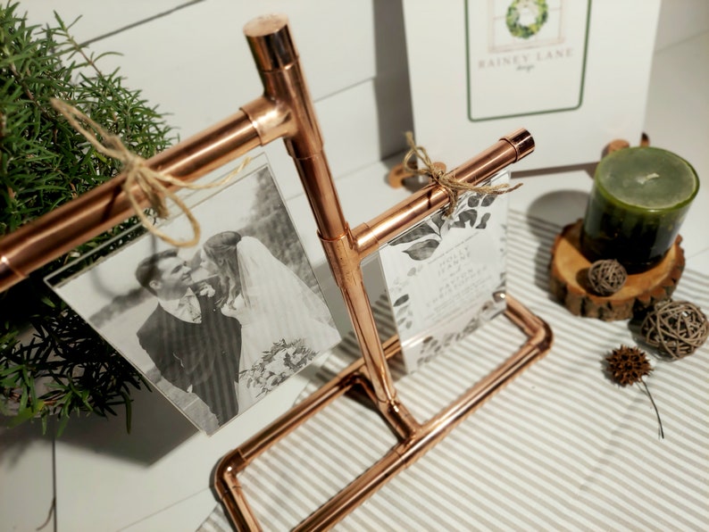 May include: Copper pipe picture frame with two photos and a wedding invitation. The frame is on a striped surface with a candle and other decorations.