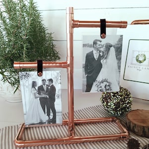 May include: Copper pipe photo display stand with three black leather straps holding up black and white wedding photos. The stand is on a striped surface with a small wooden log and pine cones.