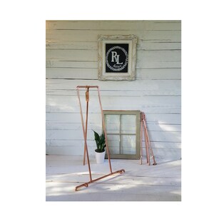 Könnte beinhalten: Ein kupferfarbener Kleiderständer mit minimalistischem Design steht vor einer weiß gestrichenen Holzwand. Der Ständer hat einen rechteckigen Rahmen und eine Querstange zum Aufhängen von Kleidung. Ein gerahmtes Schild mit den Buchstaben "RL" ist sichtbar.