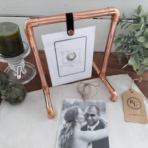 Puede incluir: Marco de fotos de tubería de cobre con una correa de cuero negro y una tarjeta blanca con el texto "Rainy Lane Designs". El marco contiene una foto en blanco y negro de una pareja.