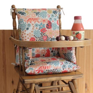May include: A vintage wooden high chair with a floral patterned cushion and backrest. A small tray holds a bottle of juice and a few wooden toys. The chair is made of light brown wood.
