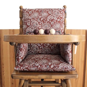 Pode incluir: Uma cadeira alta de madeira com um acolchoado estampado floral vermelho e branco. A cadeira tem uma bandeja com uma fila de contas de madeira.