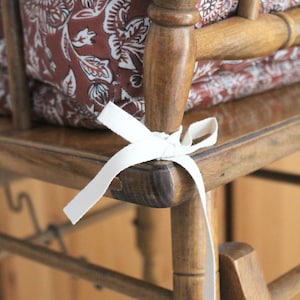 Pode incluir: Uma cadeira alta de madeira com um almofada de padr&atilde;o floral marrom e la&ccedil;os de tecido branco. A cadeira &eacute; feita de madeira natural e tem um design simples.