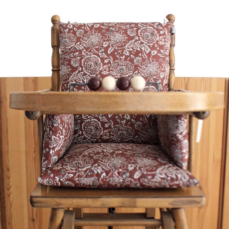 Pode incluir: Uma cadeira alta de madeira com um acolchoado estampado floral vermelho e branco. A cadeira tem uma bandeja com uma fila de contas de madeira.