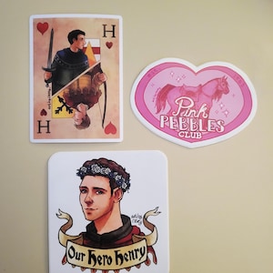 May include: Three stickers: a playing card design with a man holding a sword, a pink heart-shaped sticker with a horse, and a square sticker with a man wearing a flower crown and the text "Our Hero Henry".