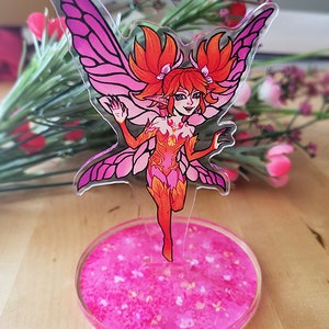 May include: A pink and orange fairy figurine with wings and a pink glitter base. The fairy has red hair and is wearing a pink and orange dress.