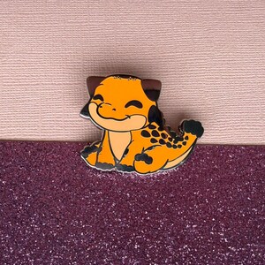 May include: A cartoon dinosaur pin in orange and black. The pin depicts a smiling dinosaur with black spots on its back and tail, brown ears, and black detailing. It is set against a pink and purple background, with a glittery lower section.