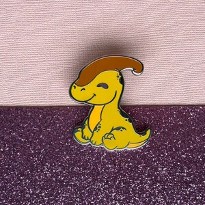 May include: A yellow dinosaur enamel pin with a brown crest and black detailing. The pin features a smiling face and is in a seated position. Set against a pink and purple glitter background.