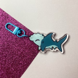 Könnte beinhalten: Ein blauer und weißer Hammerhai-Schlüsselanhänger mit einem blauen Metallschlüsselring. Der Hai ist im Cartoon-Stil gehalten und hat einen flachen, hammerförmigen Kopf.