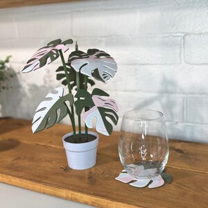 May include: A decorative paper plant with pink, white, and green leaves in a small white pot. A clear wine glass and a matching leaf-shaped coaster sit on a wooden surface. The background features a white brick wall.