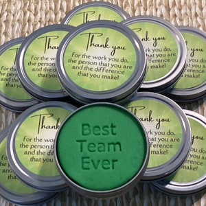 Puede incluir: Una colecci&oacute;n de peque&ntilde;as latas redondas verdes y plateadas con un mensaje de agradecimiento y las palabras "Best Team Ever" en una de las latas.