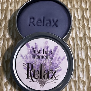 May include: A round tin container with a lavender-themed label that reads "Just for a moment... Relax". The lid is open, revealing a dark purple, scented play dough with the word "Relax" embossed on the surface.