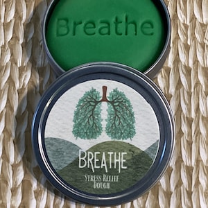 May include: A green stress relief dough in a round metal tin. The dough is embossed with the word "Breathe". The tin's lid features a watercolor illustration of lungs and the words "Breathe Stress Relief Dough".