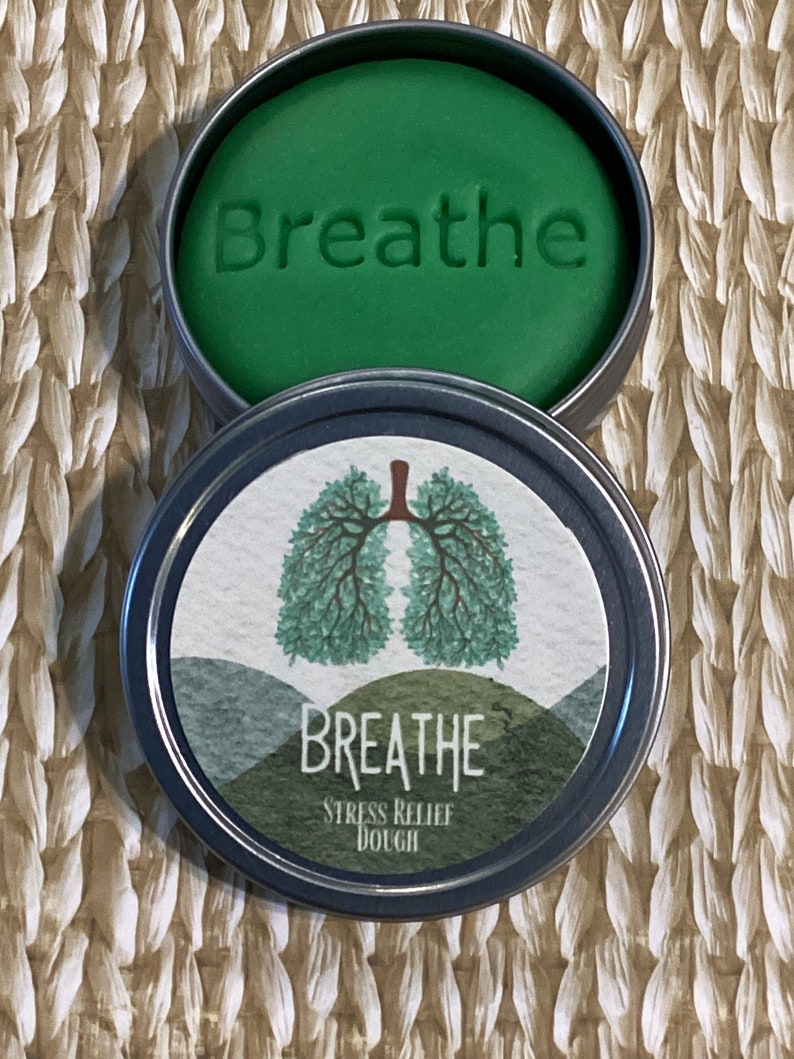 May include: A green stress relief dough in a round metal tin. The dough is embossed with the word "Breathe". The tin's lid features a watercolor illustration of lungs and the words "Breathe Stress Relief Dough".