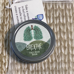 May include: A silver tin of "Breathe Stress Relief Dough" with a green lung design and the word "Breathe" in white. A small card with text is attached to a clear bag. The card reads "Take a moment to feel refreshed, breathe to relax, create a moment."