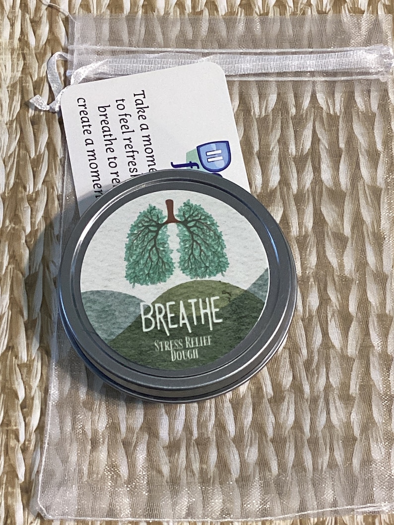 May include: A silver tin of "Breathe Stress Relief Dough" with a green lung design and the word "Breathe" in white. A small card with text is attached to a clear bag. The card reads "Take a moment to feel refreshed, breathe to relax, create a moment."
