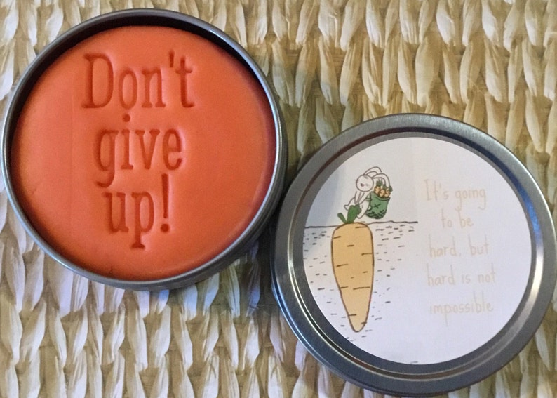 May include: A round orange tin with the text "Don't give up!" on the lid. The bottom of the tin has a white label with a cartoon rabbit carrying a basket of carrots and the text "It's going to be hard, but hard is not impossible."