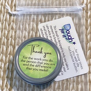 Puede incluir: Una peque&ntilde;a lata redonda de plata con una tapa verde que dice "Gracias por el trabajo que haces, la persona que eres y la diferencia que haces!" La lata est&aacute; sentada en una bolsa de malla blanca con una tarjeta blanca que dice "Dough for it...para esos momentos en los que deseas que la vida tenga un bot&oacute;n de pausa."
