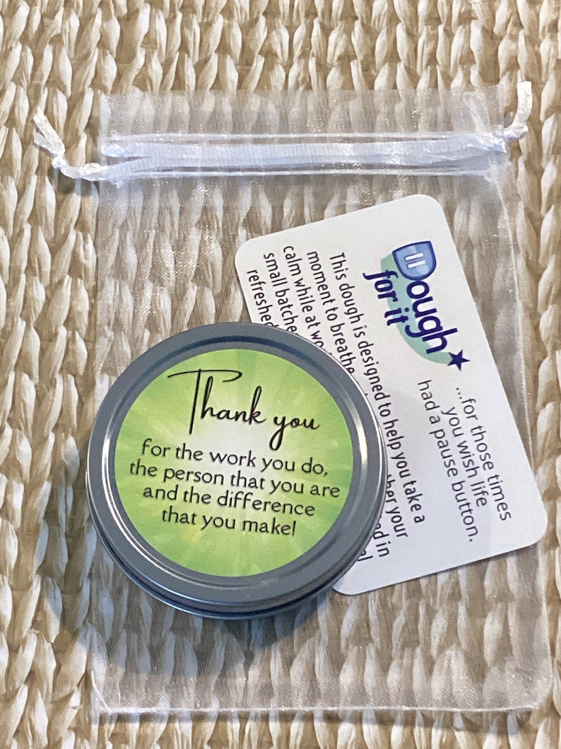 Puede incluir: Una peque&ntilde;a lata redonda de plata con una tapa verde que dice "Gracias por el trabajo que haces, la persona que eres y la diferencia que haces!" La lata est&aacute; sentada en una bolsa de malla blanca con una tarjeta blanca que dice "Dough for it...para esos momentos en los que deseas que la vida tenga un bot&oacute;n de pausa."