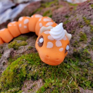 May include: A segmented orange and white toy snake with a white swirl on its head. The snake has brown eyes and is lying on a bed of green moss.